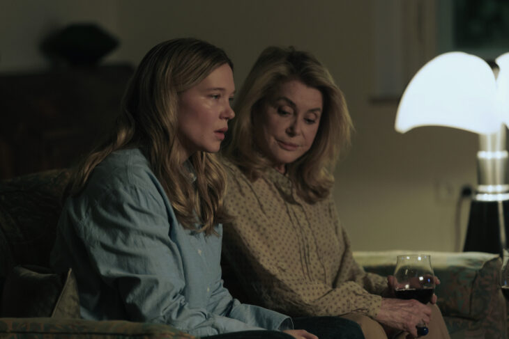 "Two women sit on a couch in a dimly lit living room, one holding a glass of wine, both appearing thoughtful or serious, with a modern lamp providing soft light in the background."